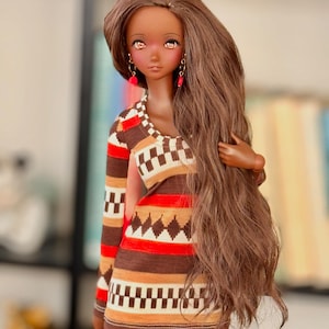 May include: A doll with long brown hair wearing a brown, red, and white patterned dress with long sleeves. The doll has brown eyes and is wearing pink earrings.