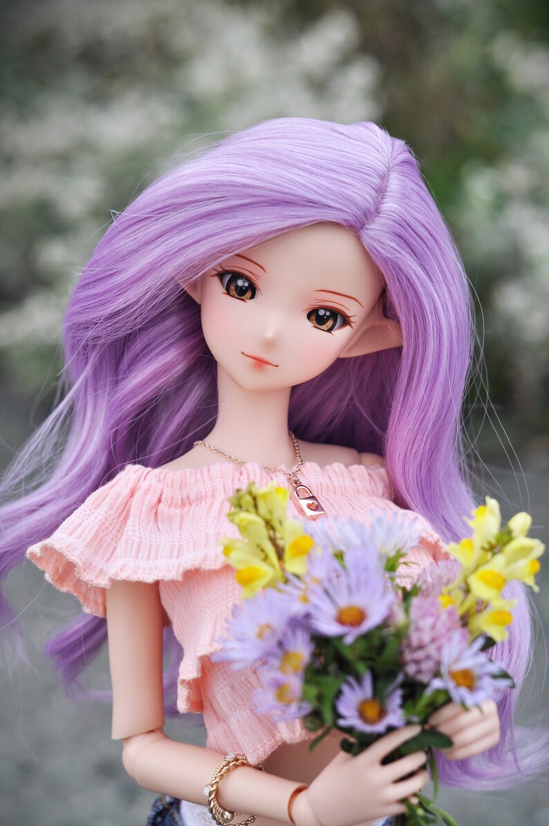 Op de afbeelding: Een pop met lang, paars haar die een roze top met blote schouders draagt en een boeket paarse en gele bloemen vasthoudt.