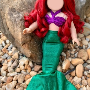 Puede incluir: Una mu&ntilde;eca de pelo rojo con un disfraz de sirena morado y verde. La mu&ntilde;eca tiene una cola verde con un acabado brillante.