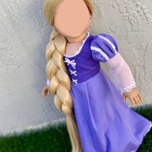 May include: A doll dressed in a purple dress with a bodice and long blonde braided hair. The dress has white lace trim and puffed sleeves. The doll is standing on green grass.
