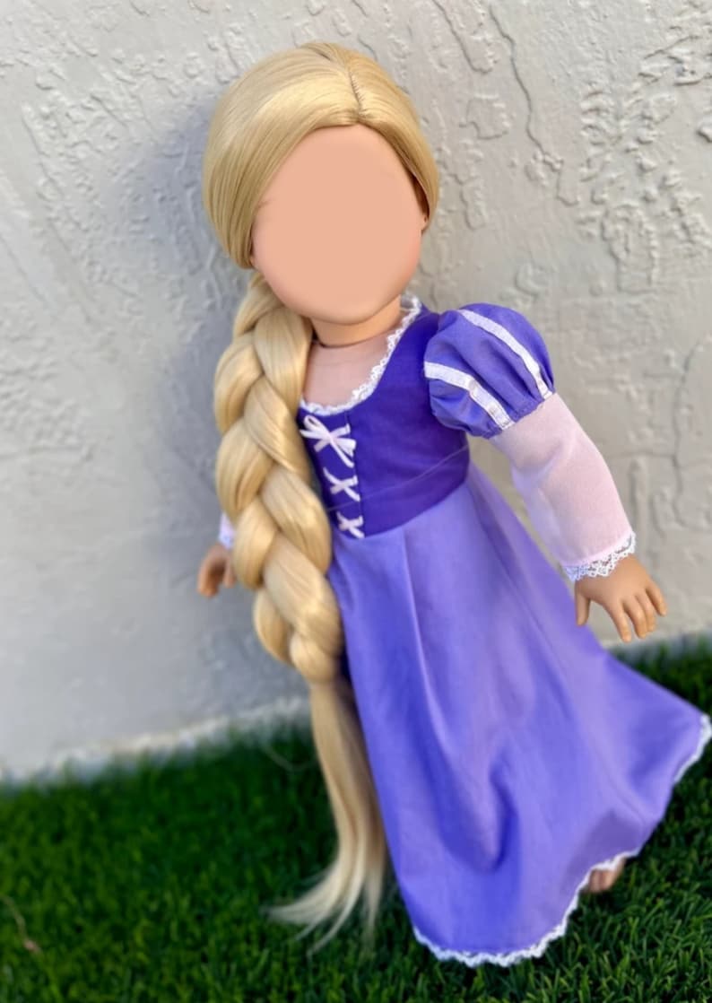 May include: A doll dressed in a purple dress with a bodice and long blonde braided hair. The dress has white lace trim and puffed sleeves. The doll is standing on green grass.