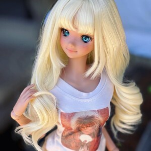 May include: A blonde doll with blue eyes wearing a white t-shirt with a graphic print of a person with long hair. The doll is wearing brown pants and has a long, wavy hairstyle.