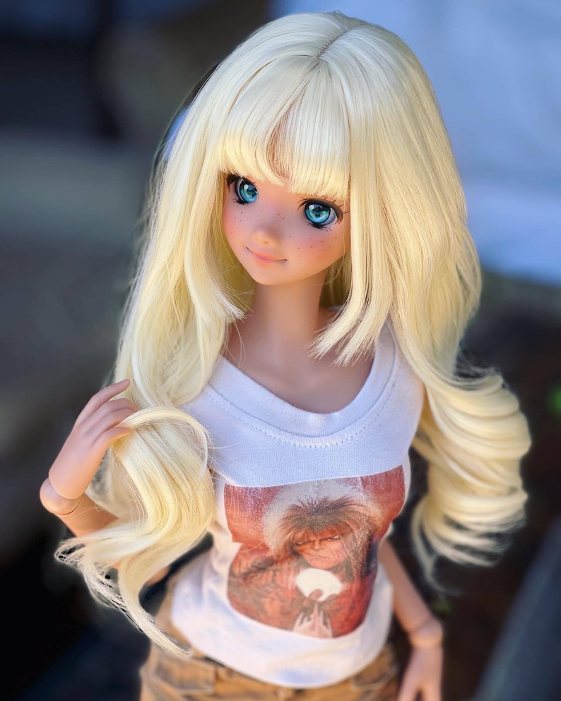 May include: A blonde doll with blue eyes wearing a white t-shirt with a graphic print of a person with long hair. The doll is wearing brown pants and has a long, wavy hairstyle.