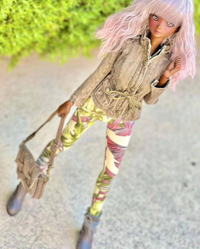 May include: A doll wearing a tan jacket, patterned leggings, and gray boots, holding a matching purse. The leggings feature a colorful floral design with green, pink, and white hues. The doll has pink hair.