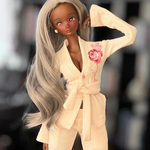 May include: A doll with long, silver hair wearing a light pink, long-sleeved,  robe with a floral design. The robe has a sash that ties at the waist. The doll has brown skin and brown eyes.