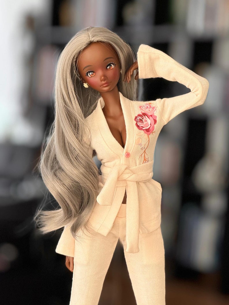 May include: A doll with long, silver hair wearing a light pink, long-sleeved,  robe with a floral design. The robe has a sash that ties at the waist. The doll has brown skin and brown eyes.