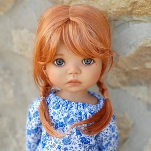May include: A doll with long, straight, auburn hair styled in two braids. The doll is wearing a blue and white floral dress with a ruffled neckline. The doll has blue eyes and a soft, innocent expression.