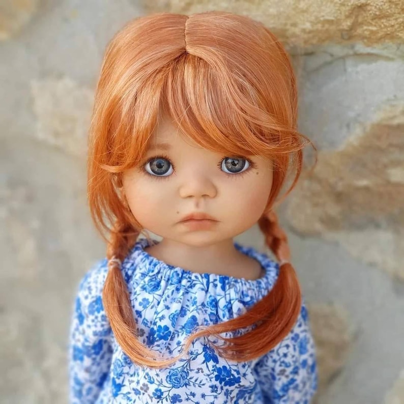 May include: A doll with long, straight, auburn hair styled in two braids. The doll is wearing a blue and white floral dress with a ruffled neckline. The doll has blue eyes and a soft, innocent expression.