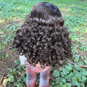 May include: A doll with dark brown, curly hair. The doll is wearing a light gray top and plaid pants in shades of red, pink, and gray. The doll is standing in a grassy area.