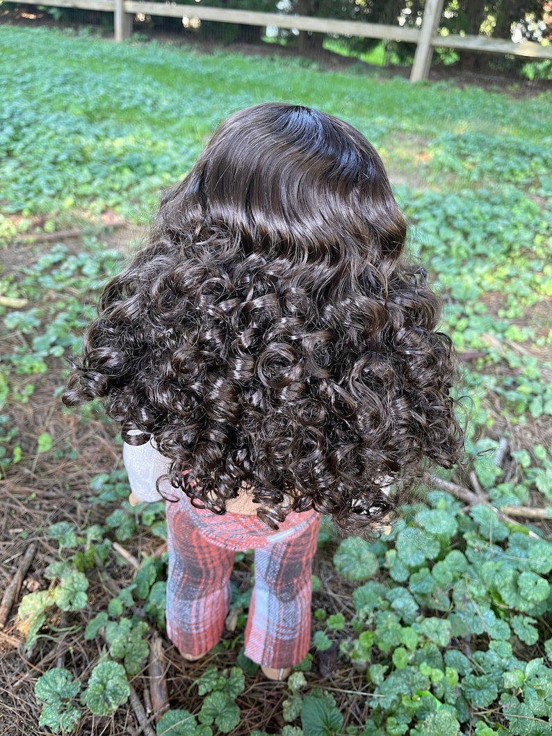 May include: A doll with dark brown, curly hair. The doll is wearing a light gray top and plaid pants in shades of red, pink, and gray. The doll is standing in a grassy area.