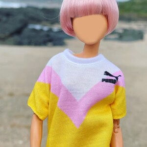 Peut inclure: Une poup&eacute;e avec des cheveux roses portant un t-shirt ray&eacute; jaune, rose et blanc avec un logo Puma noir.