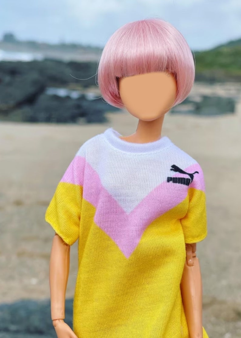 Peut inclure: Une poup&eacute;e avec des cheveux roses portant un t-shirt ray&eacute; jaune, rose et blanc avec un logo Puma noir.