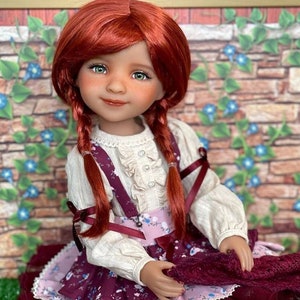 May include: A red-haired doll wearing a white blouse with a ruffled collar and a burgundy skirt with floral print. The doll has green eyes and is holding a burgundy fabric in her hands.