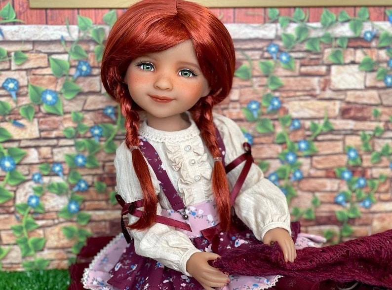 May include: A red-haired doll wearing a white blouse with a ruffled collar and a burgundy skirt with floral print. The doll has green eyes and is holding a burgundy fabric in her hands.