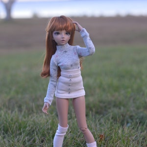 May include: A doll with long, red hair wearing a grey sweater with white snowflakes and a white skirt with buttons. The doll is standing in a grassy field.