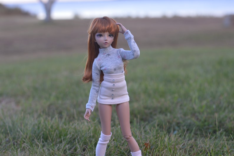 May include: A doll with long, red hair wearing a grey sweater with white snowflakes and a white skirt with buttons. The doll is standing in a grassy field.