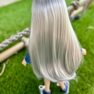 May include: A doll with long, straight, silver hair, wearing a blue denim jacket and blue sneakers. The doll is standing on green grass, with a rope and wooden structure in the background. The doll is facing away from the camera.