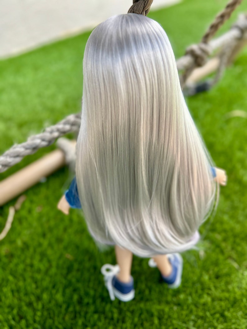 May include: A doll with long, straight, silver hair, wearing a blue denim jacket and blue sneakers. The doll is standing on green grass, with a rope and wooden structure in the background. The doll is facing away from the camera.