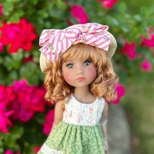 Puede incluir: Una muñeca con cabello rubio rizado y ojos verdes que lleva un vestido floral blanco y verde. La muñeca lleva un sombrero de paja con un lazo a rayas rosas y blancas. El fondo presenta flores rojas y follaje verde.