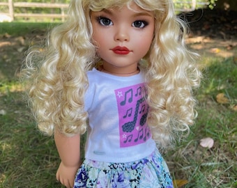 May include: A doll with blonde, curly hair, wearing a white t-shirt with musical notes and a floral skirt. The doll has red lipstick and is standing outside on the grass.