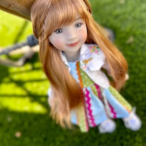 May include: A doll with red hair and blue eyes wearing a white, green and pink dress with a floral pattern. The doll is looking up at the camera.