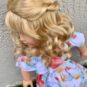 May include: A doll with long, wavy, blonde hair styled in a half-updo. The doll wears a white dress with a floral pattern in red, blue, and yellow. The dress has ruffled sleeves and a gathered waist. The doll is positioned in front of a textured gray wall and green grass.
