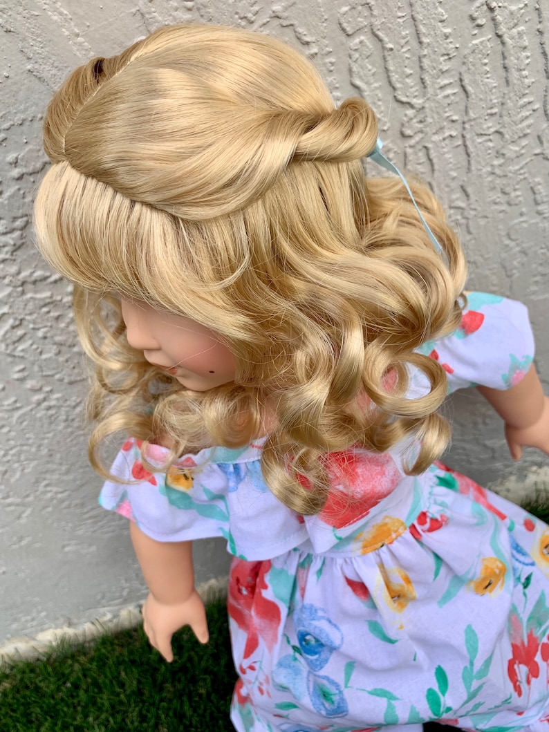 May include: A doll with long, wavy, blonde hair styled in a half-updo. The doll wears a white dress with a floral pattern in red, blue, and yellow. The dress has ruffled sleeves and a gathered waist. The doll is positioned in front of a textured gray wall and green grass.