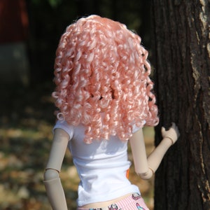 May include: A doll with long, curly, pink hair wearing a white t-shirt and pink and white patterned shorts.