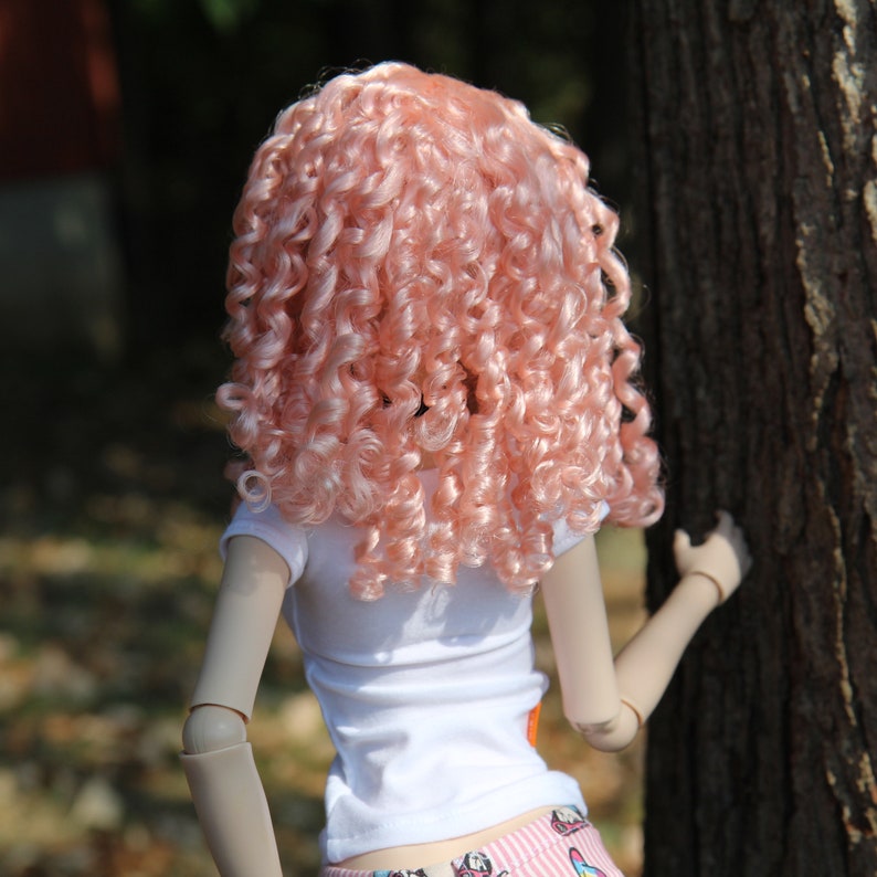 May include: A doll with long, curly, pink hair wearing a white t-shirt and pink and white patterned shorts.