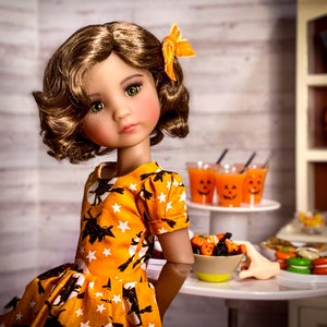 Może przedstawiać: Lalka w żółtej i pomarańczowej sukience z czarno-białym wzorem. Lalka ma brązowe włosy i zielone oczy. Lalka stoi przed stołem z dekoracjami na Halloween.