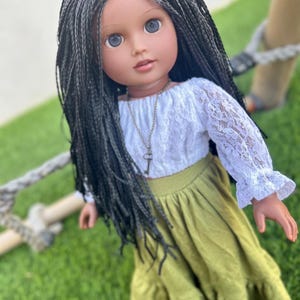 May include: A doll with long black braids, blue eyes, and a fair complexion. The doll wears a white lace top, a green skirt, and a key pendant necklace. The doll is standing on green grass.
