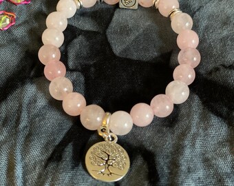 May include: Rose quartz beaded bracelet with a silver-tone tree of life charm and a silver cube bead with an Om symbol. The bracelet features light pink and white beads, strung on an elastic band.