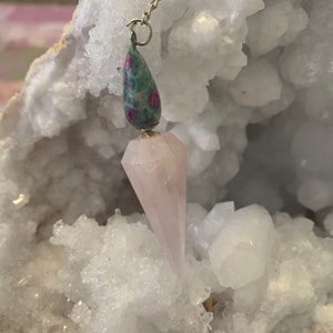 May include: A pink crystal pendulum with a green and pink stone at the top. The pendulum is hanging from a silver chain.