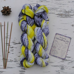 May include: Two skeins of yarn in a yellow and grey colourway. The yarn is hand-dyed and has a speckled appearance. The yarn is handmade by Echoacraftings OY. The yarn is made with alpaca, merino, cotton, bamboo, linen, and silk. The yarn is machine washable and can be tumble dried on a low setting.