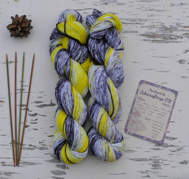 May include: Two skeins of yarn in a yellow and grey colourway. The yarn is hand-dyed and has a speckled appearance. The yarn is handmade by Echoacraftings OY. The yarn is made with alpaca, merino, cotton, bamboo, linen, and silk. The yarn is machine washable and can be tumble dried on a low setting.