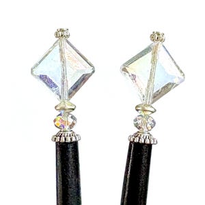 May include: Two black hair sticks with decorative tops. Each stick features a clear, iridescent, diamond-shaped crystal, a smaller faceted crystal, and ornate silver-colored metal accents. The sticks are set against a white background.