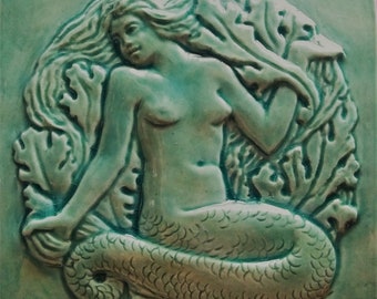 Puede incluir: Un azulejo de cerámica verde con una escultura en relieve de una sirena. La sirena está representada con el pelo suelto y una cola de pez. El azulejo está decorado con algas y corales.