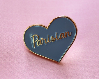 May include: A heart-shaped pin with a gold border and the word "Parisian" in gold script. The heart is a muted blue color and rests on a pink fabric background. The pin is a fashion accessory.