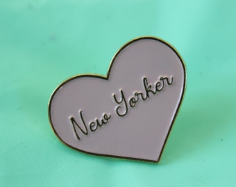 May include: A purple heart-shaped enamel pin with a gold outline. The pin says "New Yorker" in black cursive lettering.