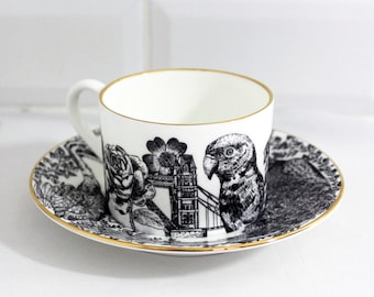 May include: A black and white teacup and saucer set with a gold rim. The design features a parrot, a rose, Tower Bridge, and other London-themed imagery.