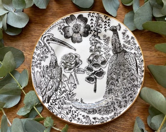 May include: A white ceramic plate with a black and white design featuring a peacock, a bird, flowers, and leaves. The plate has a gold rim.