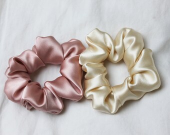 May include: Two satin scrunchies, one in a dusty rose color and the other in a cream color. The scrunchies are lying on a white surface.
