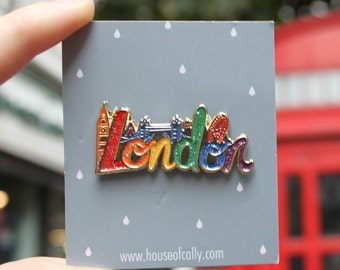 May include: A rainbow enamel pin with the word "London" in a stylized font. The pin features iconic London landmarks, including Big Ben, the London Eye, and a red double-decker bus. The pin is on a gray card with white raindrops and the website "www.houseofcally.com".
