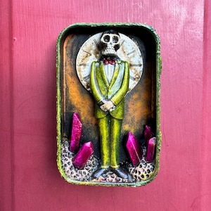 May include: A rectangular tin container featuring a skeletal figure in a green suit and red bowtie standing before a clock face background. The scene includes several bright pink crystal-like shards protruding from a textured base within the tin.