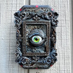 May include: A rectangular wall plaque featuring an ornate, dark-toned frame with intricate scrollwork and small red accents. Centered within the frame is a textured, stone-like surface containing a single, realistic human-like eye with a green iris.