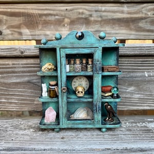 May include: A teal-colored miniature cabinet with multiple shelves, housing various curiosities. It features a small skull, bottles with cork stoppers, a clock, a seashell, and a small statue of a bird. The cabinet is decorated with small spheres and a black gem.
