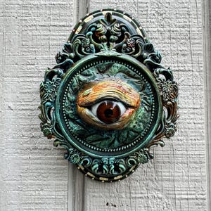 May include: A decorative wall plaque featuring a central, realistic brown eye set within an ornate, oval-shaped frame. The frame is finished in a weathered, dark teal and copper patina with intricate floral and scrollwork detailing throughout.