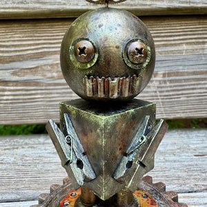May include: A steampunk metal robot sculpture with a round head, screw eyes, and a gear on top. The robot has a square body with clothespin arms and stands on a gear base. The sculpture has a weathered, aged appearance.