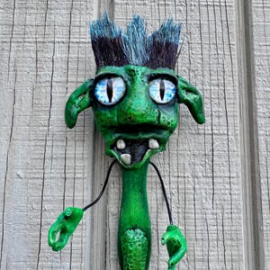 May include: A whimsical, green creature sculpture with blue-tipped hair, large blue eyes, and a toothy grin. The sculpture has outstretched arms and is mounted on a wooden surface. The creature's body is elongated and textured.