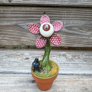 May include: A decorative sculpture featuring a flower with five red, honeycomb-textured petals and a central realistic brown eye. The flower has a green stem rising from a small terracotta pot filled with green moss and a tiny black bird figure.
